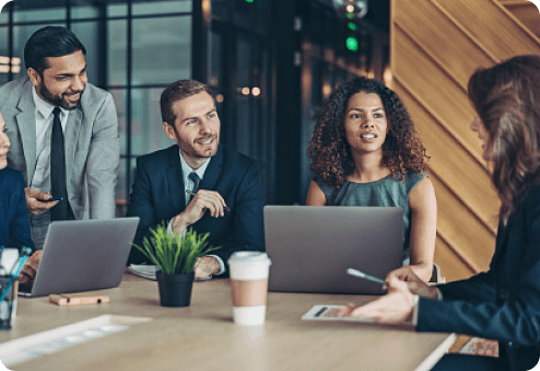 Four professionals in a meeting with laptops at a table in a modern office setting.