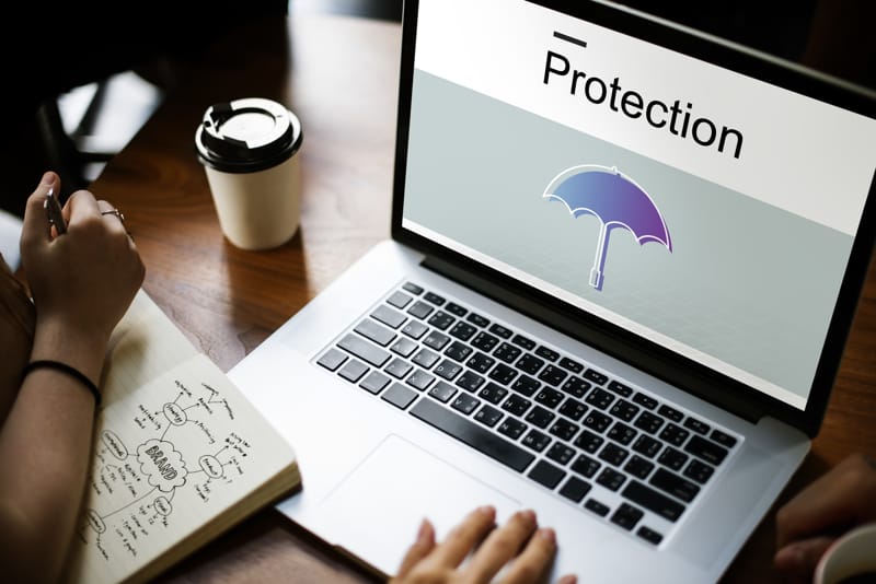 Laptop screen displaying word "Protection" with an umbrella icon, notebook beside it, and a coffee cup.