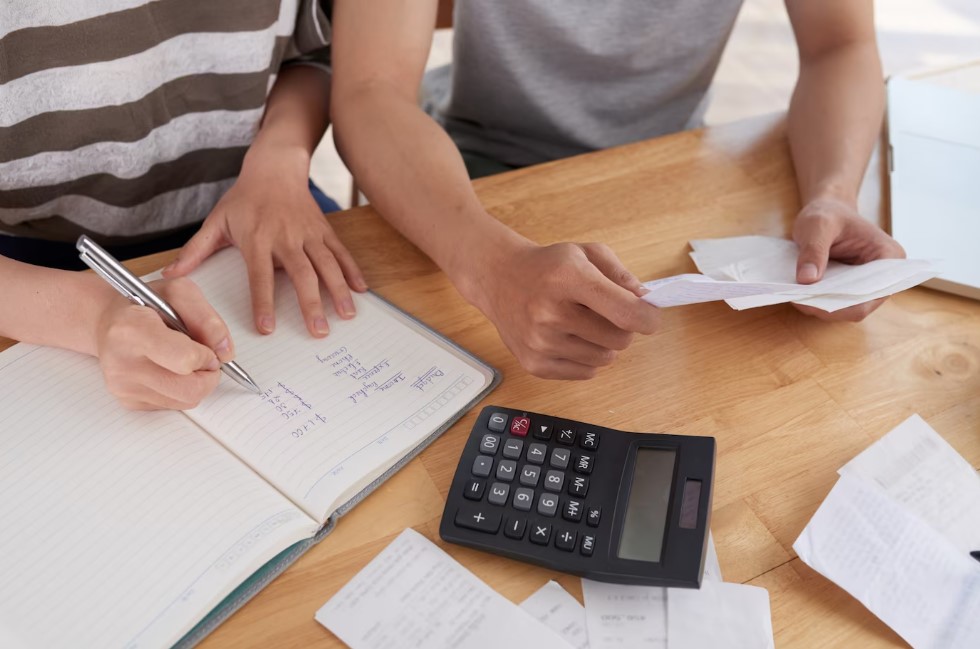 Two individuals budgeting with calculator, notebook, and receipts on a wooden table.