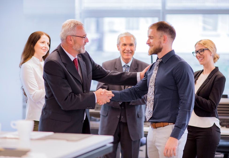 Two professionals shaking hands in an office setting, with colleagues nearby.