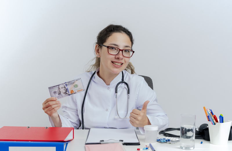 A person is holding a piece of paper indoors. They are wearing glasses and other clothing, likely related to their job. The background features a wall and a table, indicating an employment context.