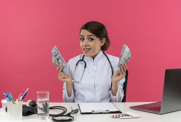 A medical professional in a white coat with a stethoscope, holding cash in both hands, at a desk with medical equipment and a laptop.