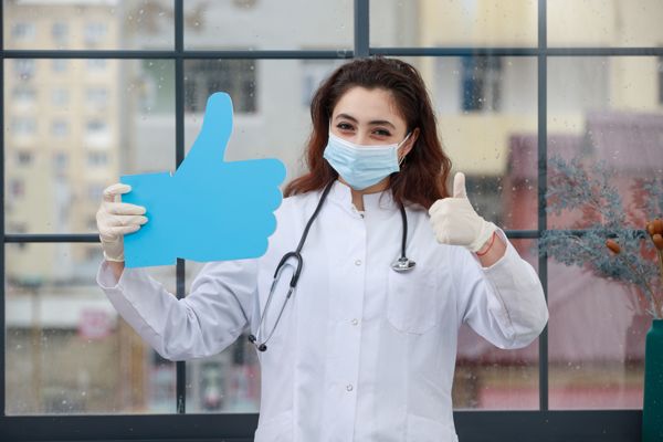 A person in a white lab coat holding a large blue thumbs-up sign and giving a thumbs up.