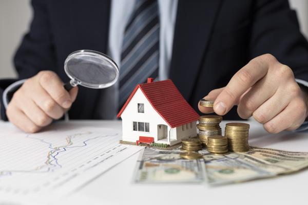 Person examining house model and coins with magnifying glass, suggesting property valuation or investment analysis.