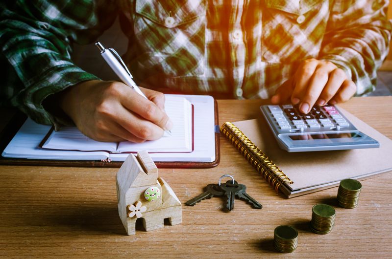 Person writing in notebook with calculator, coins, keys, and house model on desk.
