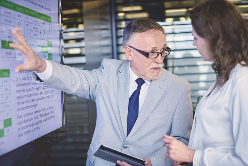 Two professionals discussing data on a digital screen, one holding a tablet.