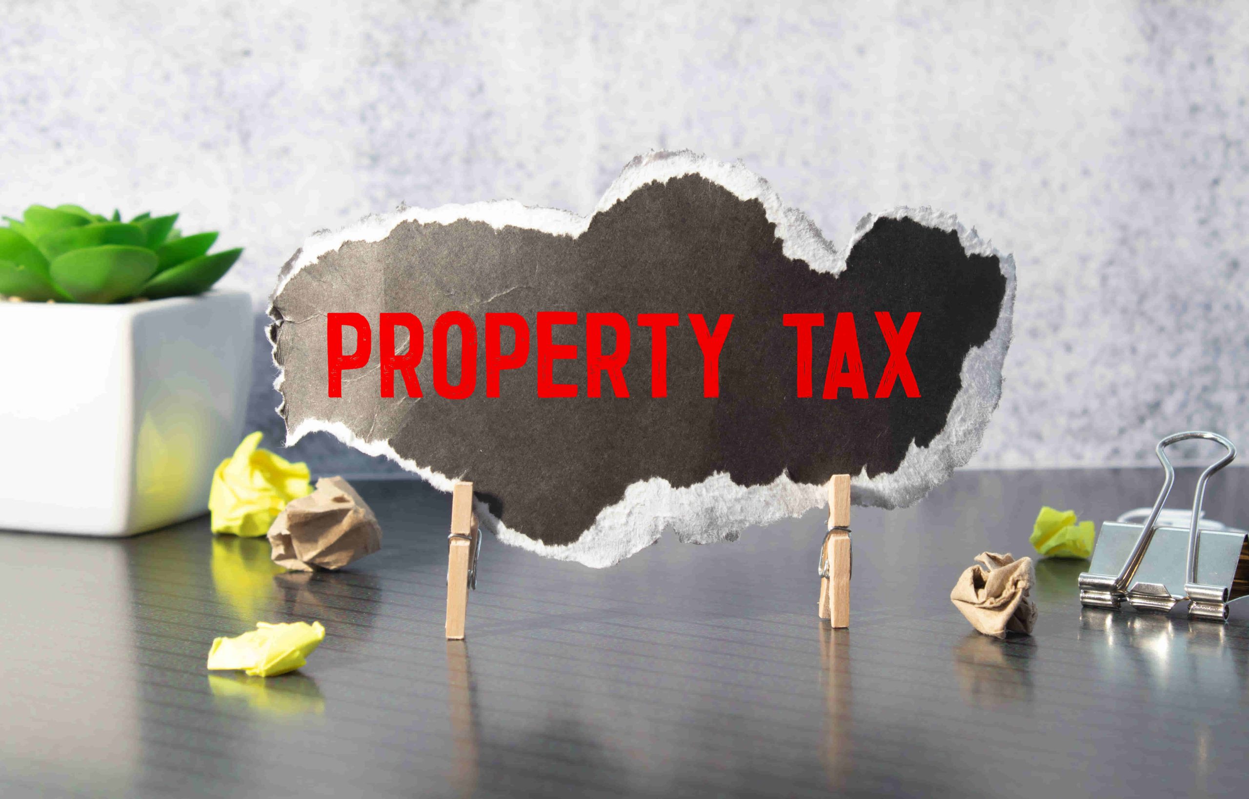 A torn paper with "PROPERTY TAX" text, held by clothespins on a desk with office supplies.