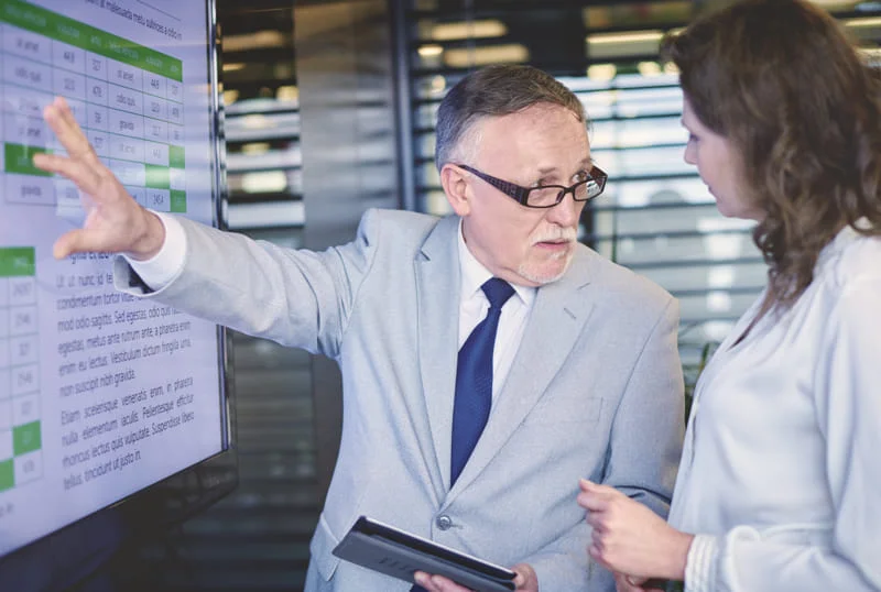 Two professionals discussing data on a digital screen, one holding a tablet.