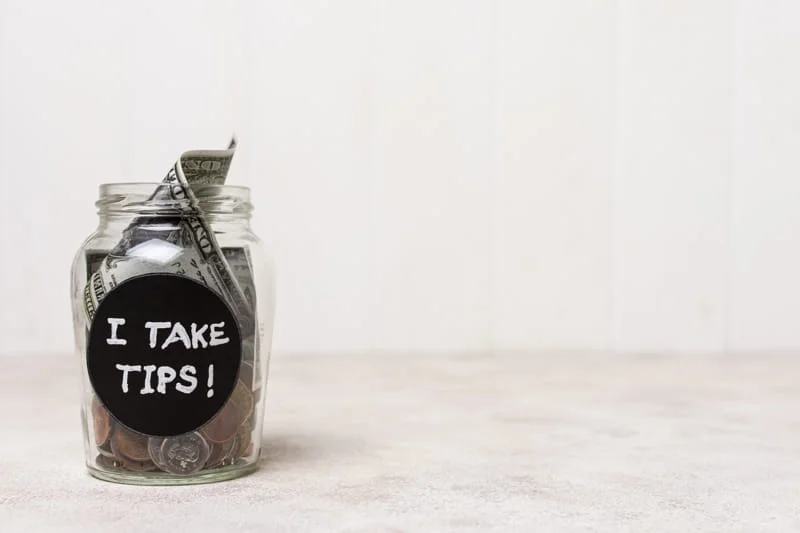 Glass jar with coins and bills labeled "I TAKE TIPS!" on light background.